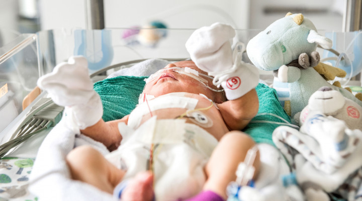 Ein Neugeborenes mit Handschuhen und Söckchen liegt in einem Bettchen, ein grosses Pflaster auf der Brust.