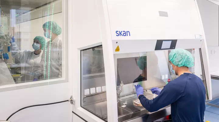 Ein Forscher sitzt vor einem Gerät und zieht etwas mit einer Spritze auf. Durch ein Fenster sieht man den angrenzenden Raum, wo sich zwei Forscherinnen befinden. Sie alle tragen Schutzkleidung, ein Haarnetz, Mundschutz und Handschuhe.