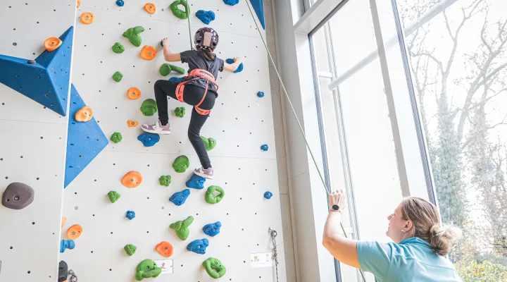 Ein Kind klettert an einer Kletterwand hoch und wird von einer Therapeutin gesichert