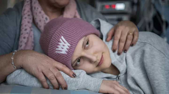 Eine Onkologiepatientin trägt eine Mütze und einen Pullover. Sie liegt auf dem Bett. Im Hintergrund sieht man den Oberkörper ihrer Mutter.
