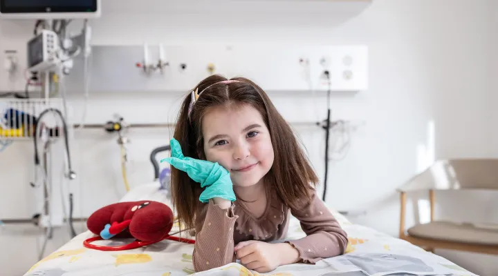 Nephrologie-Patientin Naila liegt auf einem Spitalbett im Kinderspital Zürich. Neben ihr liegt eine Plüsch-Niere und sie trägt einen grünen Gummihandschuh. Sie lächelt in die Kamera.