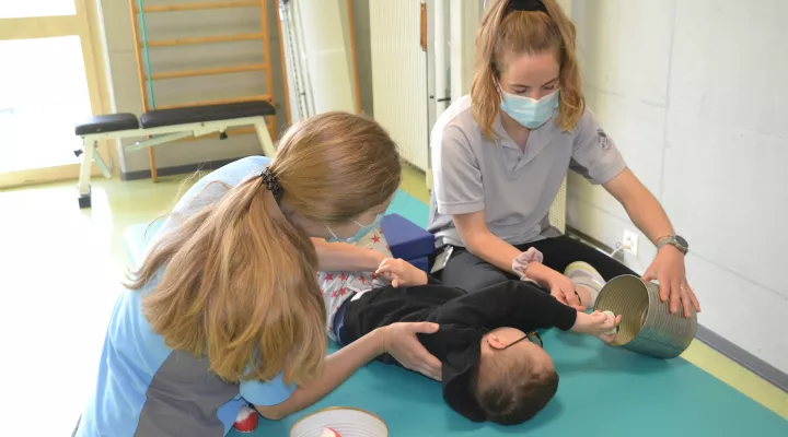 Zwei Reha-Pflegerinnen mit einem Patient, der Bälle von einer Dose in die andere wirft.