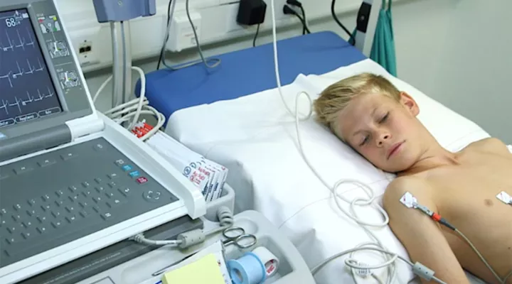 Junge oben ohne liegt auf dem Bett, neben ihm ein Monitor. Am Körper sind Drähte befestigt.