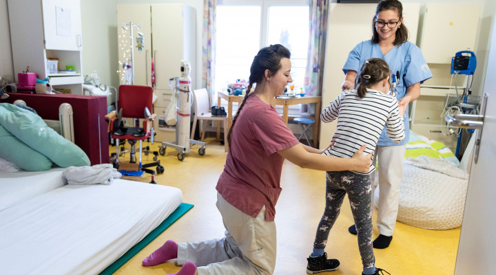 Zwei Pflegefachfrauen laufen mit einem Mädchen im Patientenzimmer in der Kinder Reha Schweiz