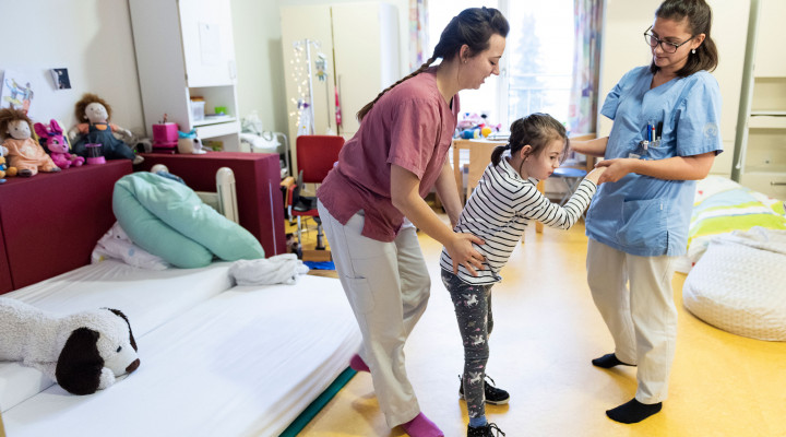 Zwei Pflegefachfrauen laufen mit einem Mädchen im Patientenzimmer in der Kinder Reha Schweiz