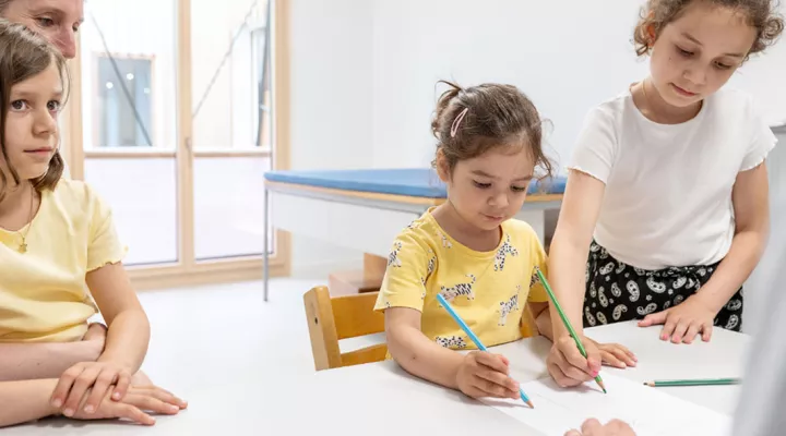 Neuropädiatrie, Neurometabolische Erkrankung, zwei Mädchen zeichnen auf ein weisses Papier, daneben sitzt Mutter und ein weiters Mädchen