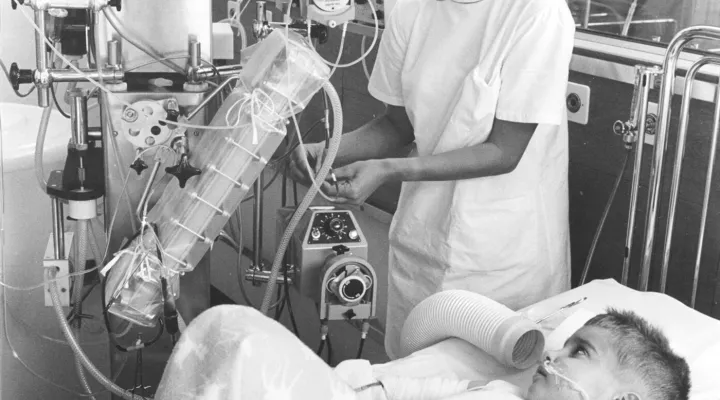 Ein Schwarzweiss-Foto, ein Kind liegt auf dem Spitalbett, angeschlossen an eine Maschine, davor steht eine Pflegefachfrau.