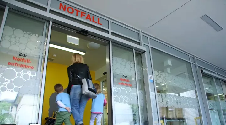 Eine Mutter hält ihre beiden Kindern an ihren Händen, man sieht sie von hinten in den Notfall hineingehen.