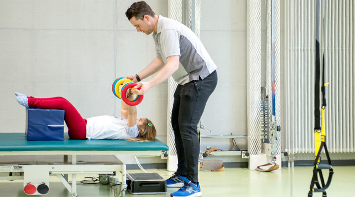 Kinder-Reha Schweiz, Sporttherapie, Patientin hebt Gewichte und Therapeut unterstützt sie dabei