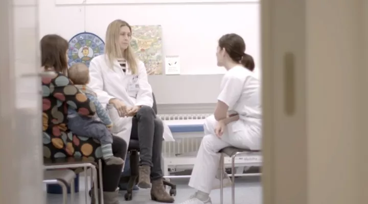 Zwei Fraun in weissen Kitteln und eine Frau mit Kleinkind sitzen in einem Sprechzimmer.