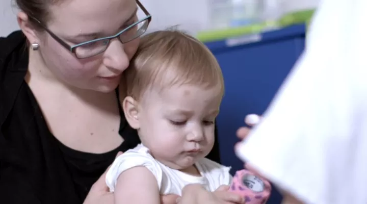 Eine Mutter mit Brille hat ein Kleinkind auf dem Schoss, im Vordergrund der weisse Ärmel einer medizinischen Person.