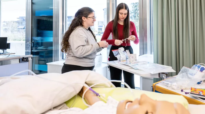 Skillslab, zwei Pflegefachfrauen in Ausbildung während des praktischen Unterrichts im Skillslab am Bett mit Puppe