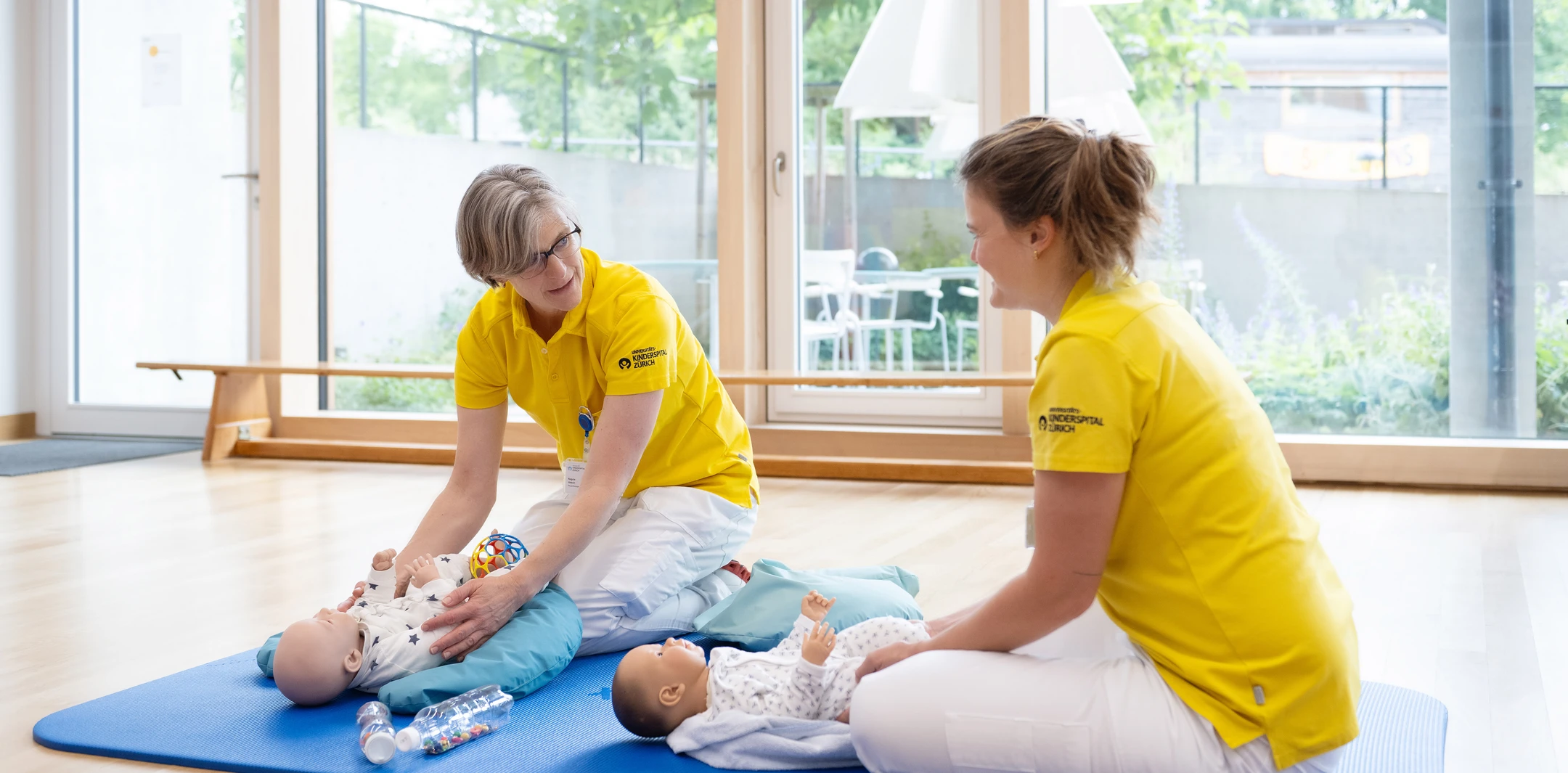 Zwei Physiotherapeutinnen sitzen auf einer blauen Matte im Therapiezentrum und demonstrieren Übungen an Babypuppen