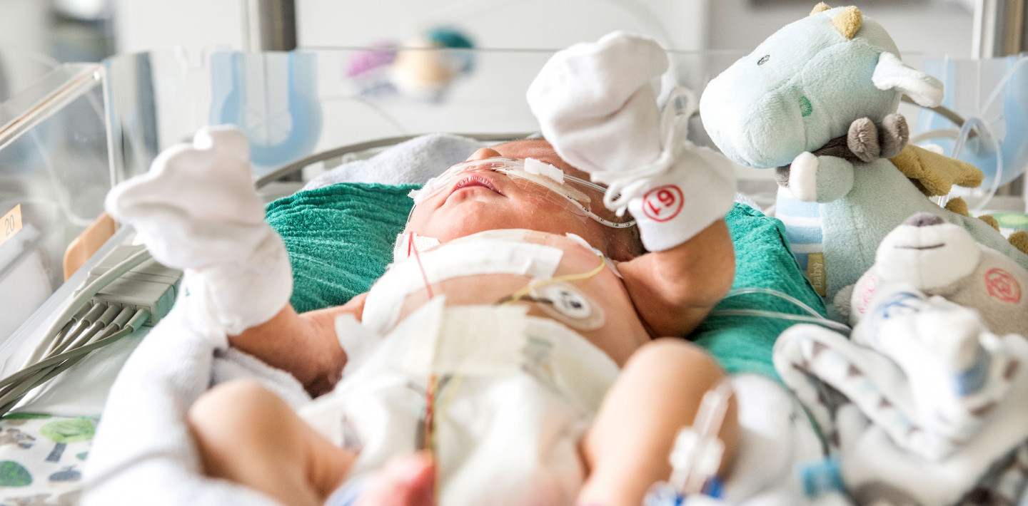 Ein Neugeborenes mit Handschuhen und Söckchen liegt in einem Bettchen, ein grosses Pflaster auf der Brust.