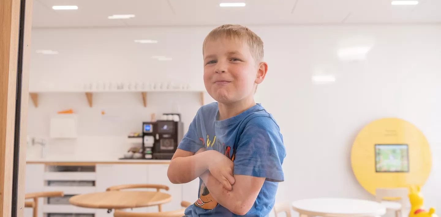 Ein Kardiologiepatient sitzt in einem Aufenthaltsraum hinter einer Glasscheibe und schaut zur Kamera. Seine Arme sind verschränkt.