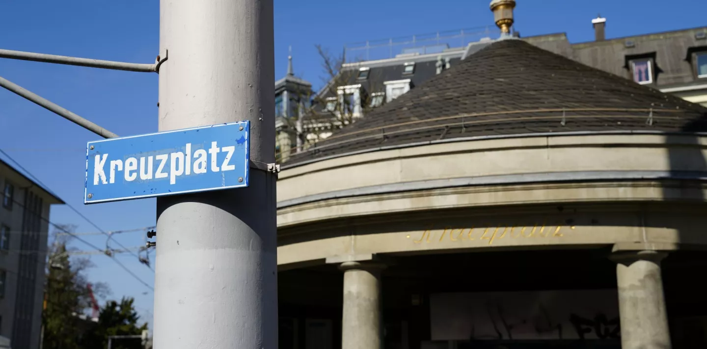 Ein Strassenschild mit der Aufschrift "Kreuzplatz" befestigt an einem Mast.