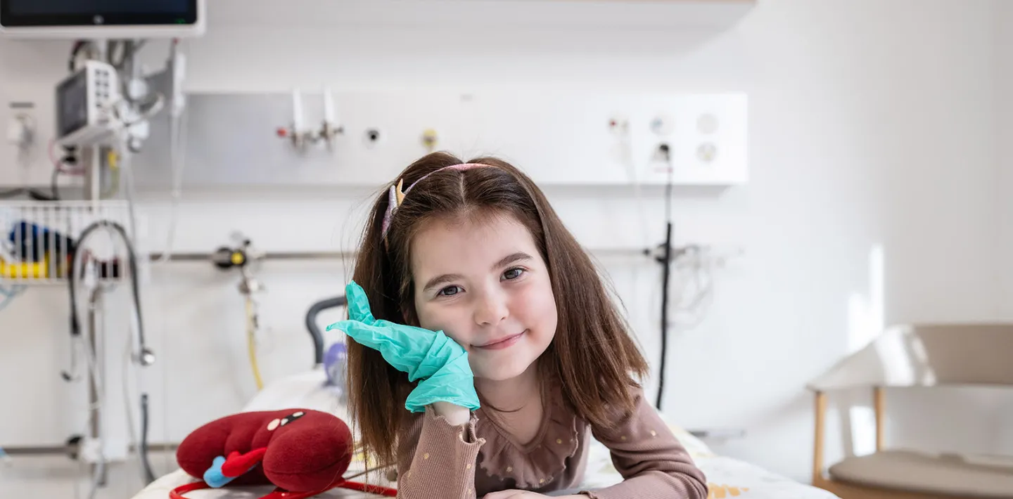 Nephrologie-Patientin Naila liegt auf einem Spitalbett im Kinderspital Zürich. Neben ihr liegt eine Plüsch-Niere und sie trägt einen grünen Gummihandschuh. Sie lächelt in die Kamera.