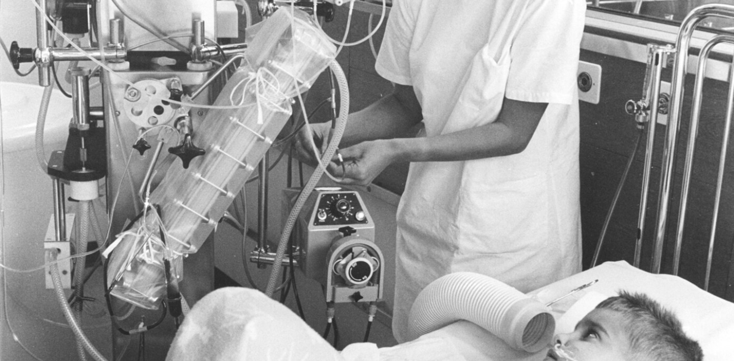Ein Schwarzweiss-Foto, ein Kind liegt auf dem Spitalbett, angeschlossen an eine Maschine, davor steht eine Pflegefachfrau.