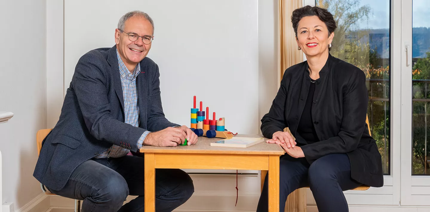 Oskar Jenni und Bea Latal sitzen an einem Kindertisch und lachen in die Kamera.