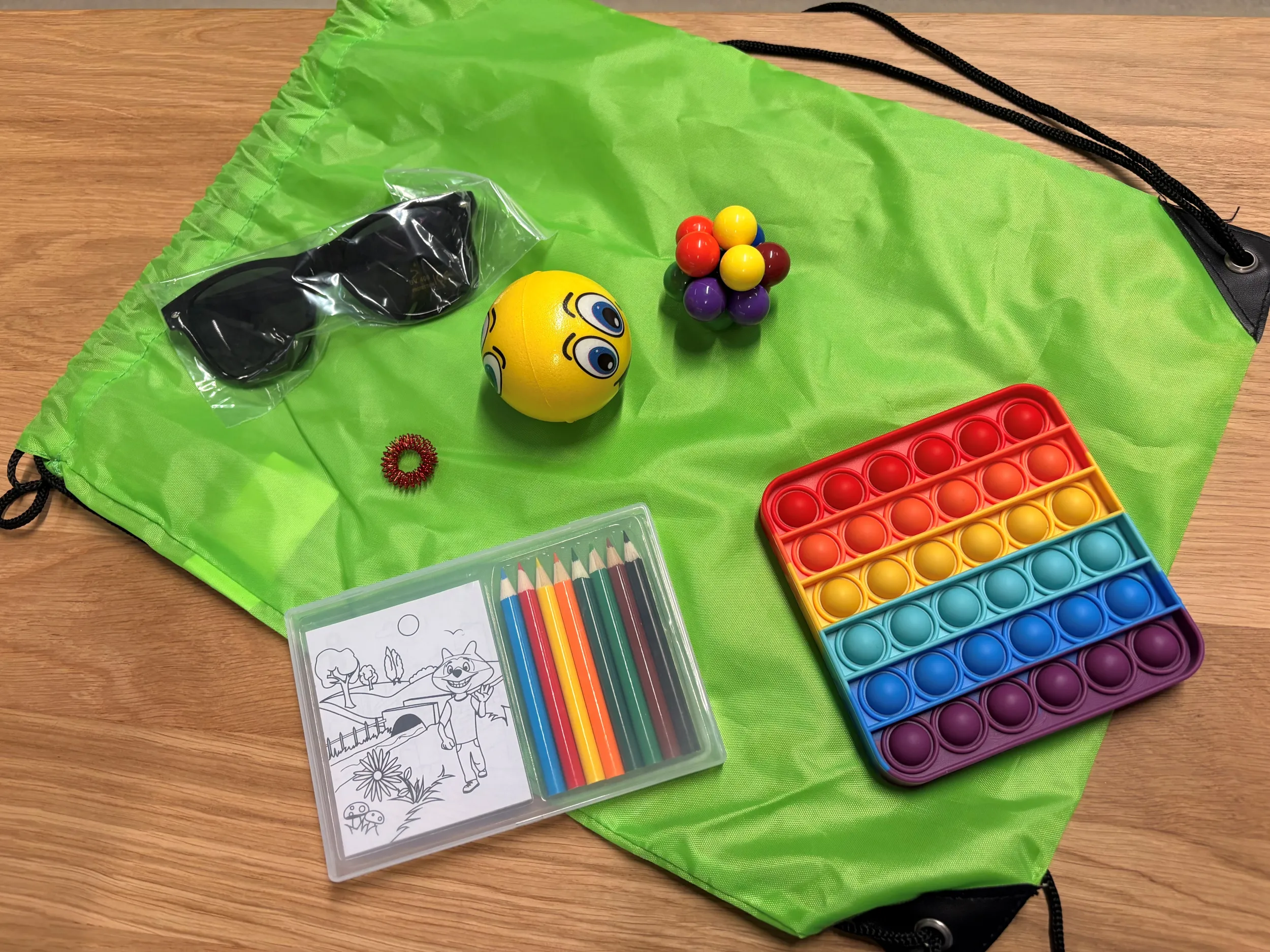 Auf einem Tisch liegen eine grüne Tasche mit verschiedenen Materialien wie Sonnenbrille, Ohropax, Fidget Toys, Ringe und weiteres Spielzeug für neurodivergente Personen
