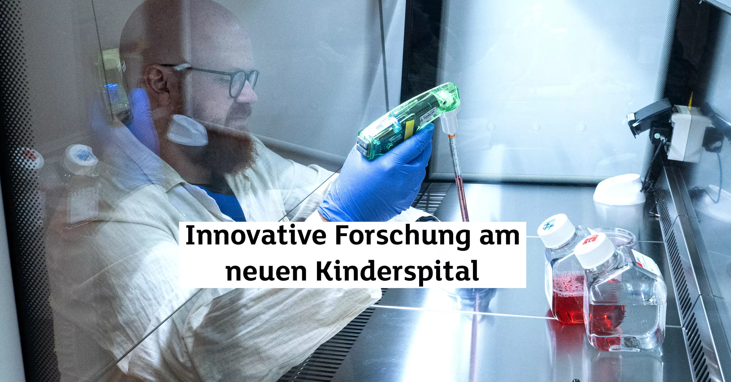 Ein Forscher mit Brille und Vollbart trägt einen weissen Kittel. Er sitzt hinter einer Glaswand und arbeitet mit Proben.
