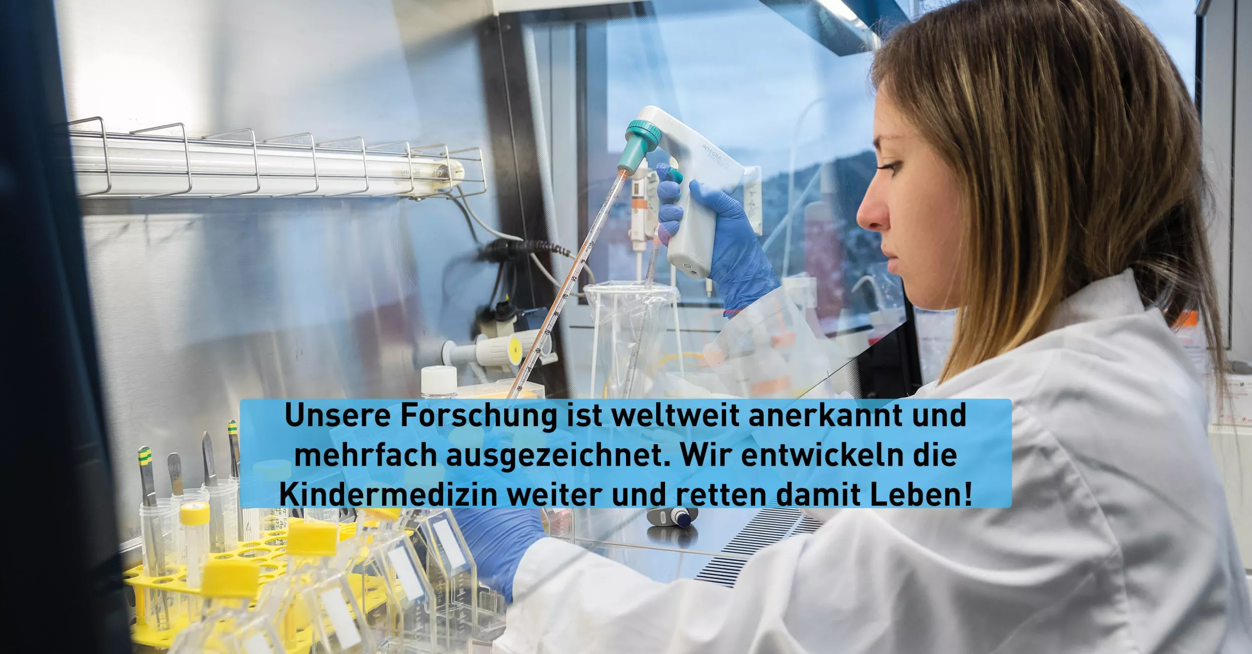 Eine Forscherin mit braunen Haaren und weissem Kittel sitzt vor einer Glaswand. Hinter der Glaswand befinden sich Ablagen mit Reagenzgläsern.