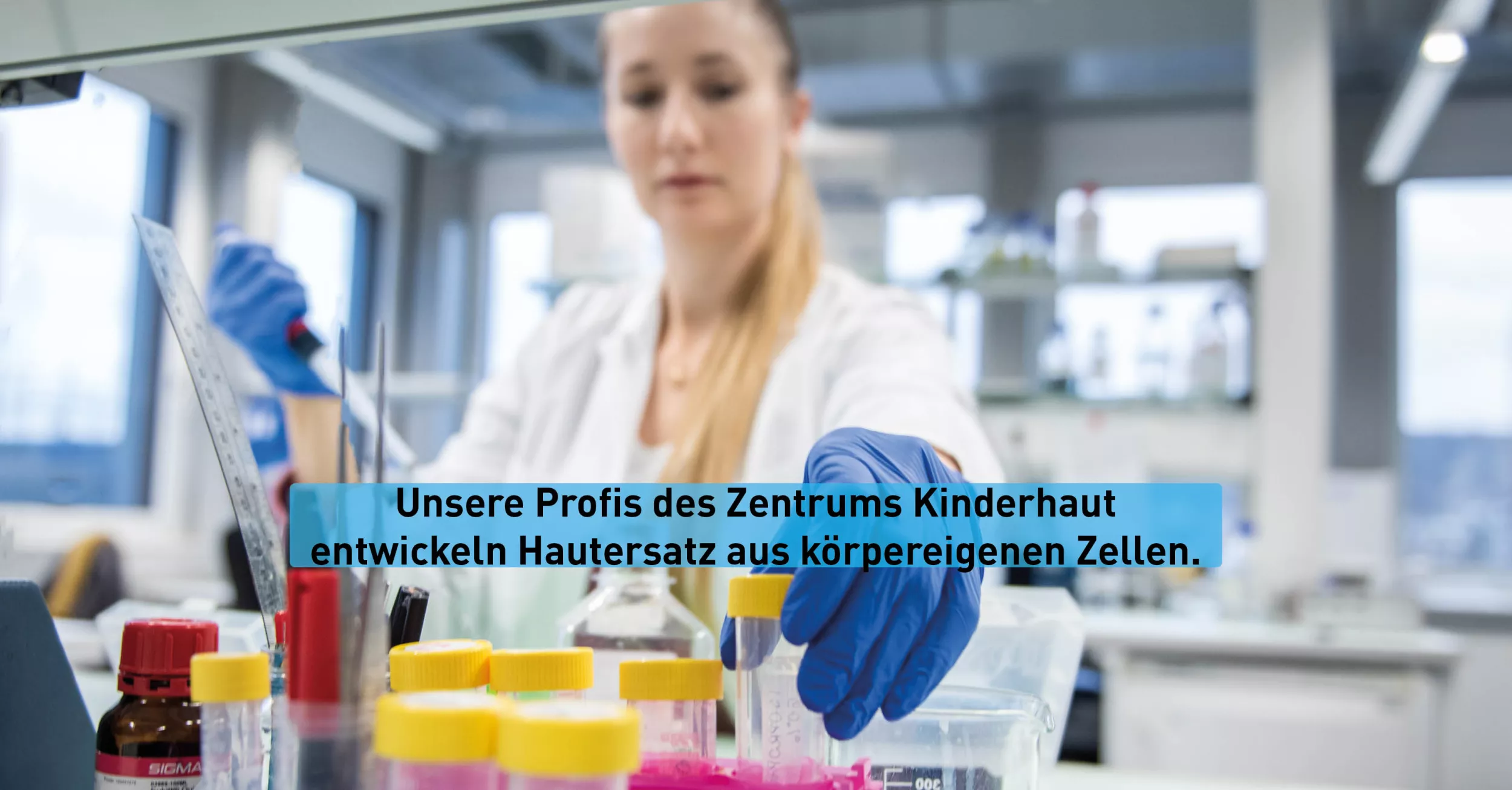 Im Vordergrund sieht man verschiedene Probengläser. Im Hintergrund ist eine blonde Forscherin. Sie trägt einen Kittel und Handschuhe.