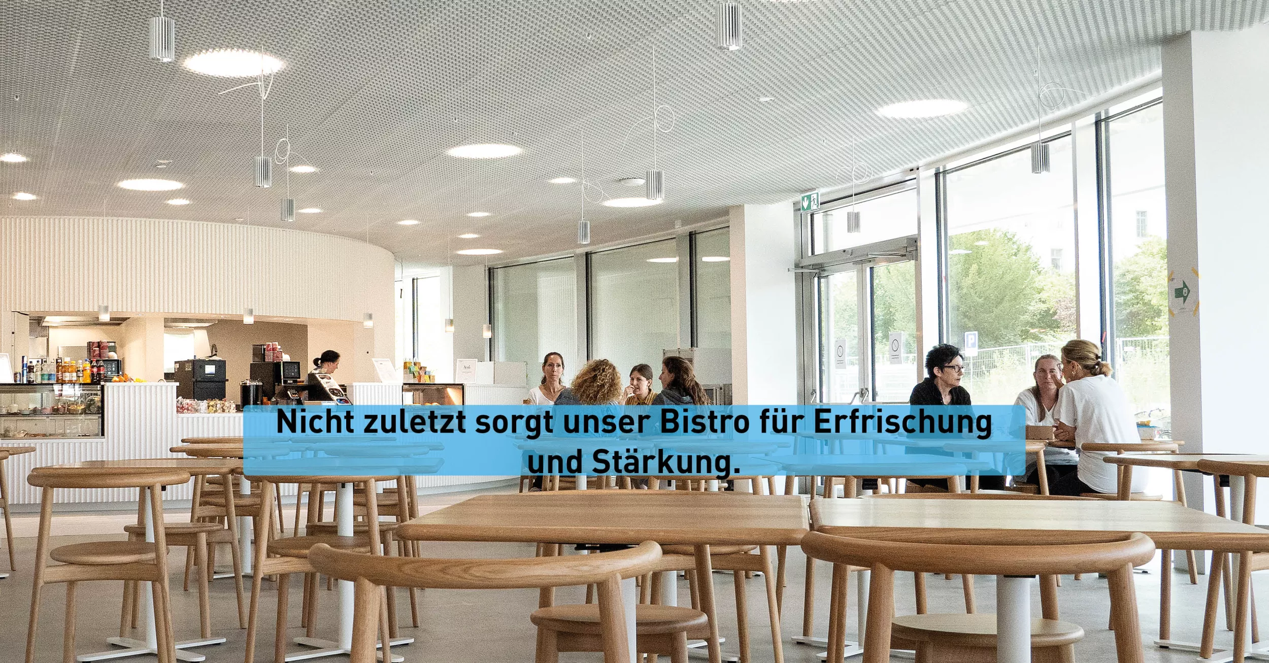Im Bistro stehen mehrere Holzstühle und Tische. Einige Personen sitzen im Bistro und unterhalten sich. Im Hintergrund sieht man einen Ausschnitt des Bistros.