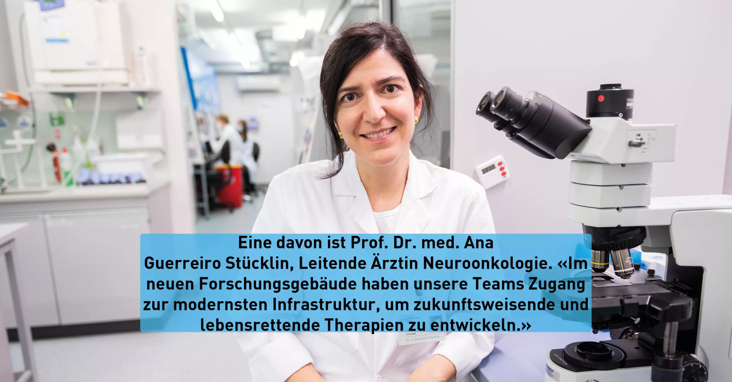 Eine dunkelhaarige Frau trägt einen Kittel und sitzt. Rechts von ihr ist ein Mikroskop. 