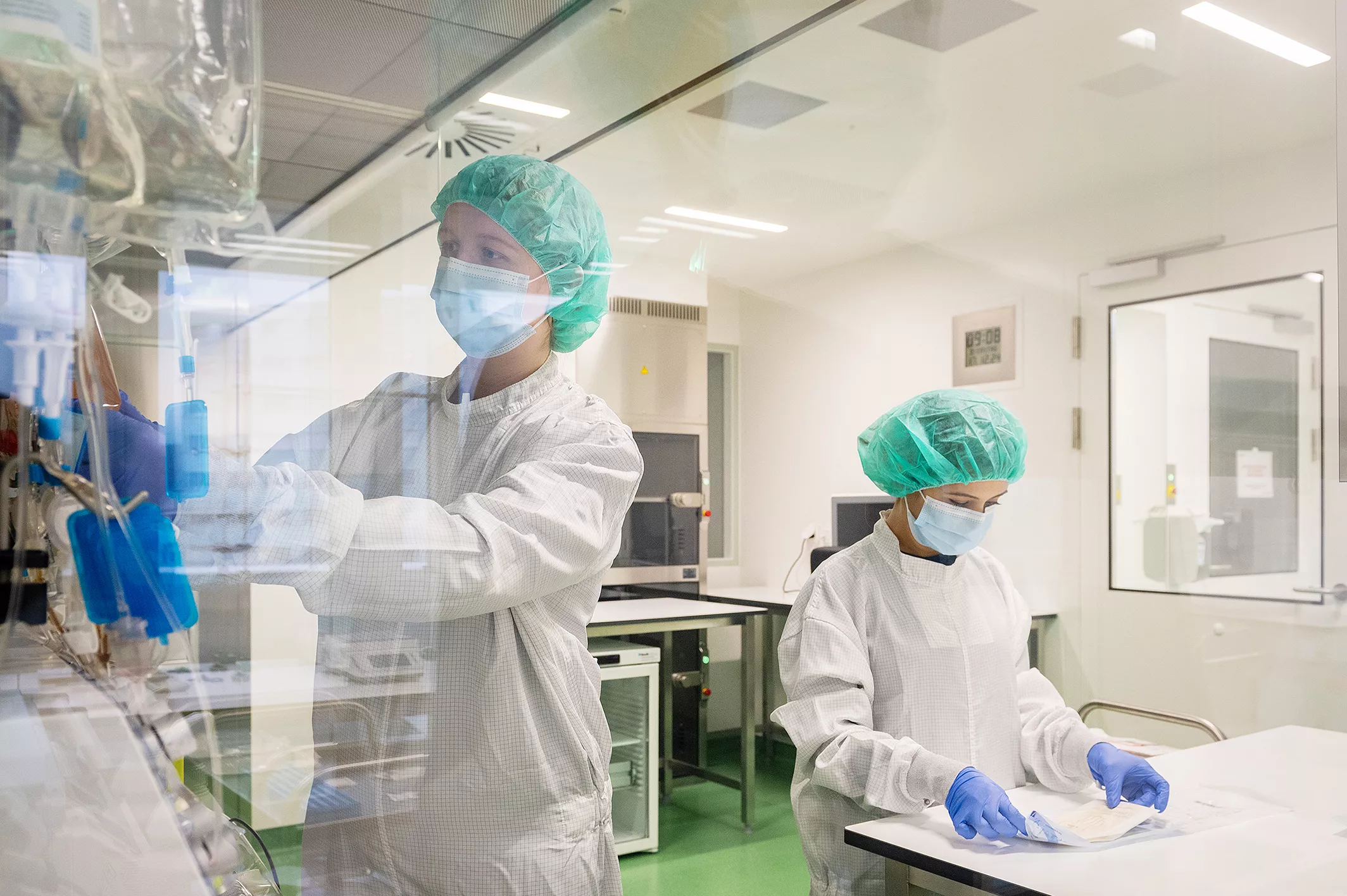Zwei Forscherinnen stehen in einem Labor. Sie tragen Schutzkleidung, ein Haarnetz, Mundschutz und Handschuhe.