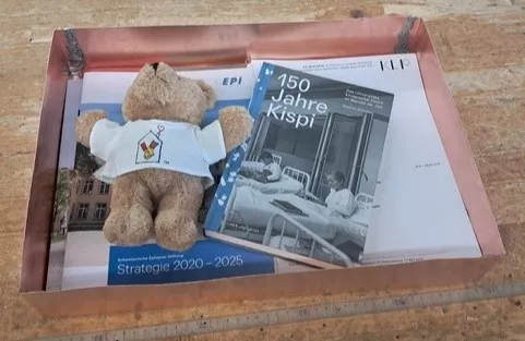 Eine Box, die als Zeitkapsel dient, liegt auf dem Boden. In der Box sieht man einen Teddybären, ein Buch sowie diverse Dokumente. 