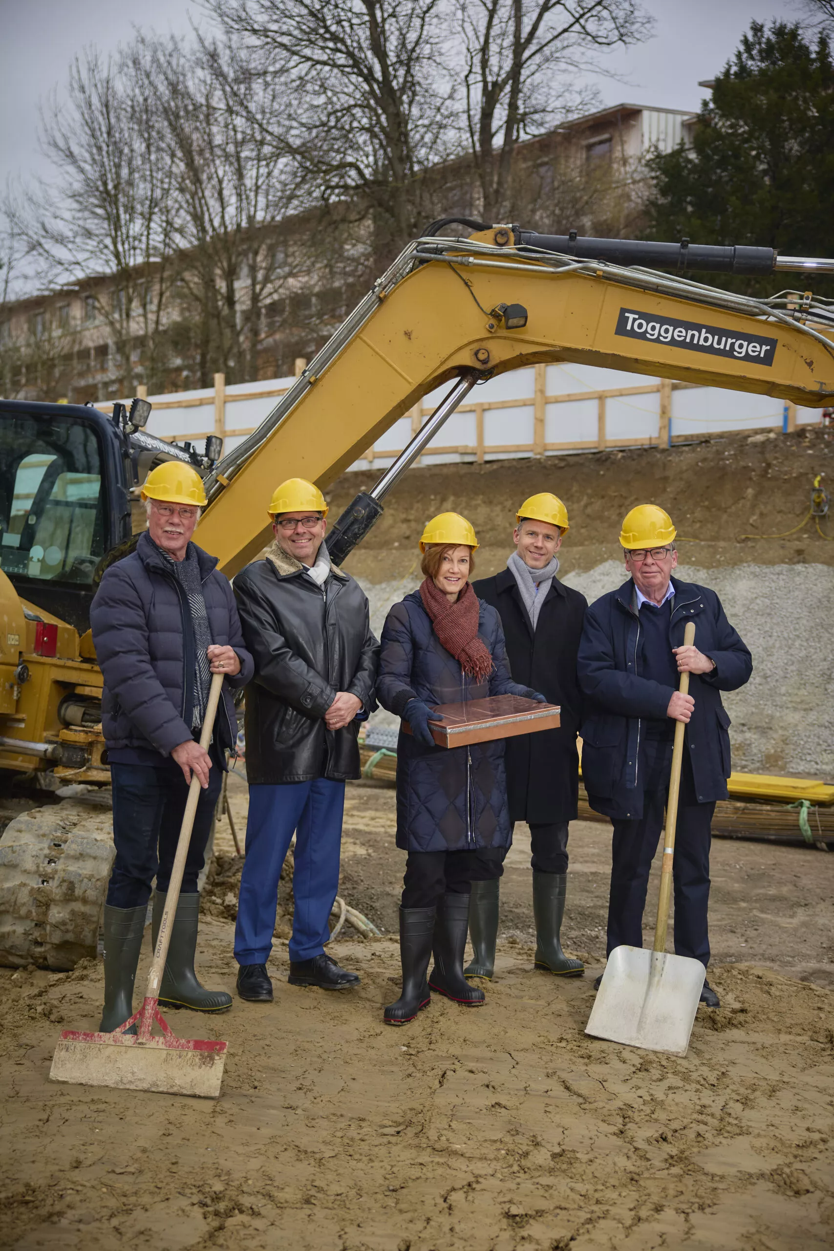 Grundsteinlegung: Thomas Schneiter Stiftungsratspräsident RMHC, Marco Beng CEO Epilepsie-Stiftung (EPI), Andrea Hemmi Direktorin RMHC, Jürg Syz Stiftungsrat EPI, Martin Vollenwyder Stiftungsratspräsident Eleonorenstiftung stehen in der Baugrube vor einem Bagger.