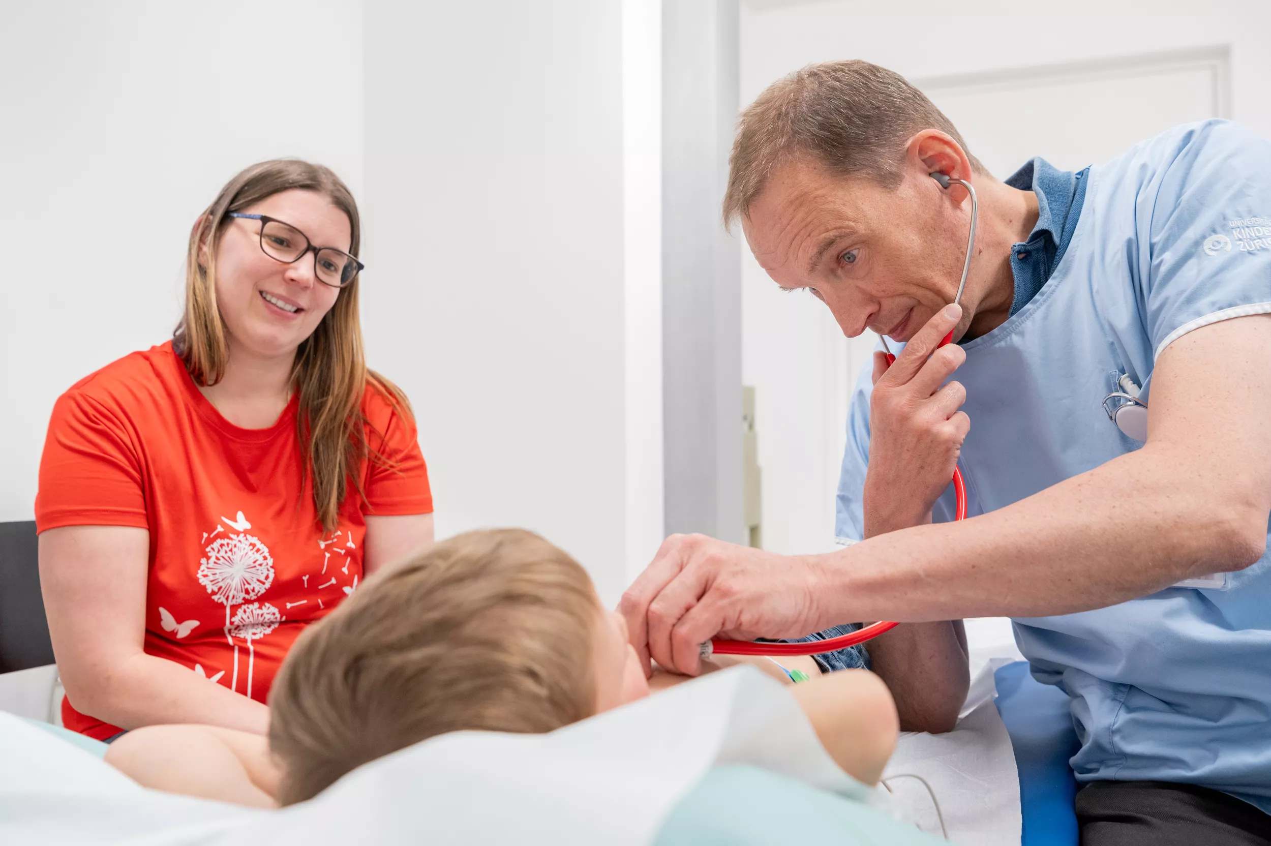 Ein Kardiologiepatient ist auf einer Behandlungsliege. Neben ihm sitzen sein Kardiologe und seine Mutter. Der Kardiologe hört den Patienten mit einem Stethoskop ab.