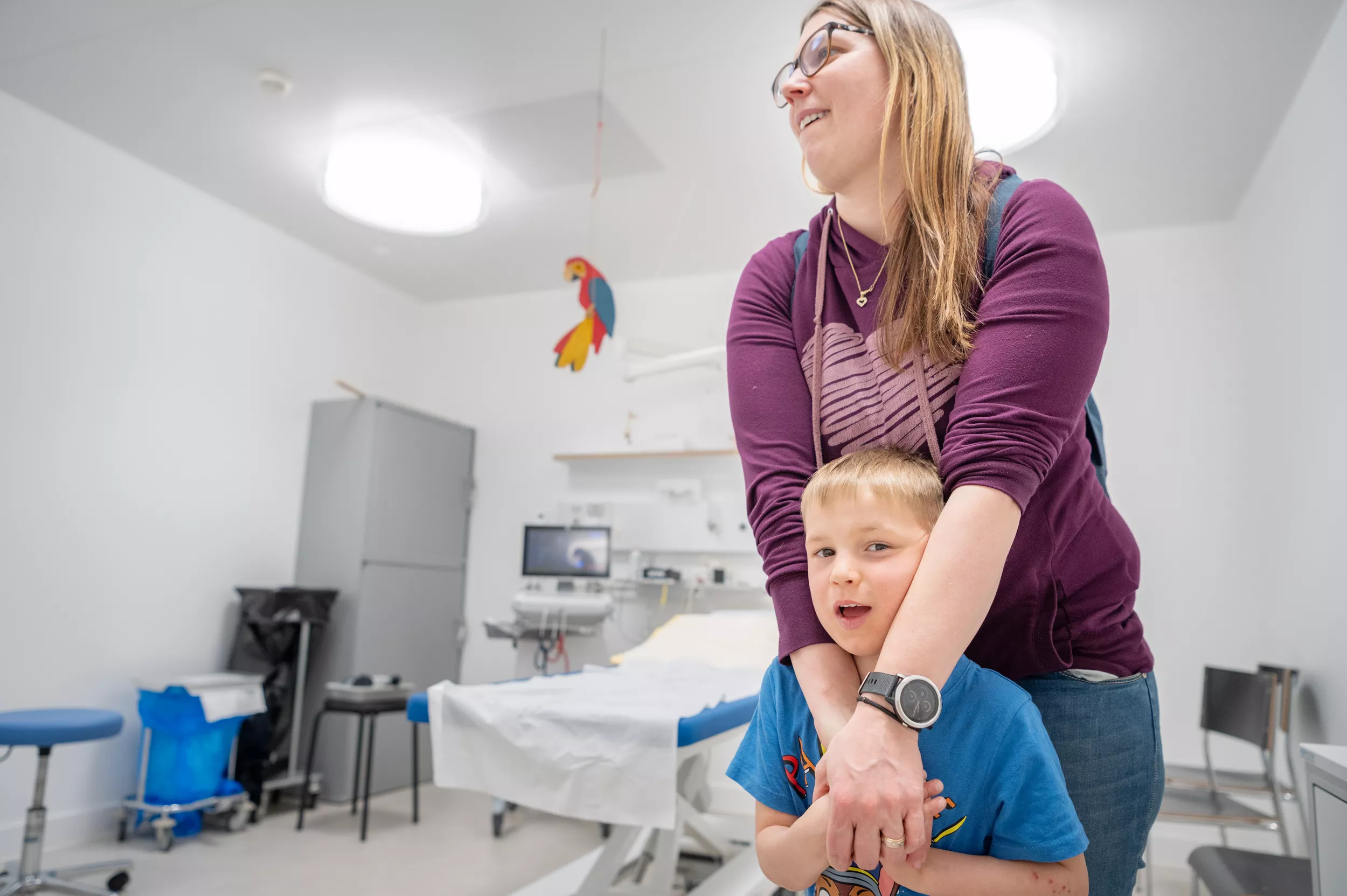 Ein Kardiologiepatient steht in einem Behandlungszimmer. Seine Mutter steht hinter ihm und hält eine Hand von ihm.