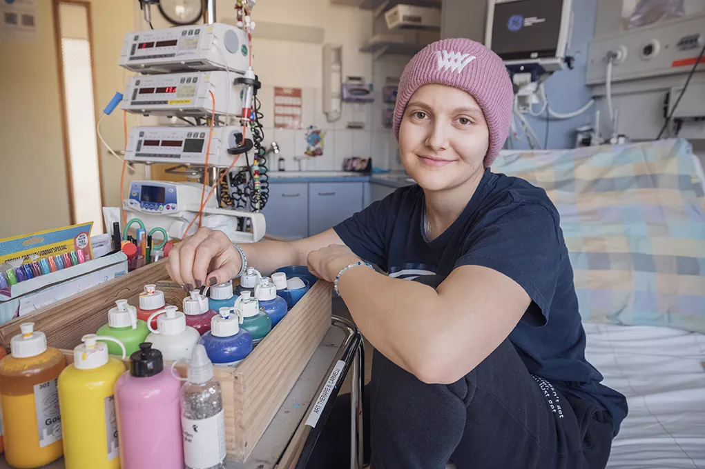 Eine junge Patientin sitzt auf einem Bett mit einer Box mit bunten Farbflaschen und Stiften