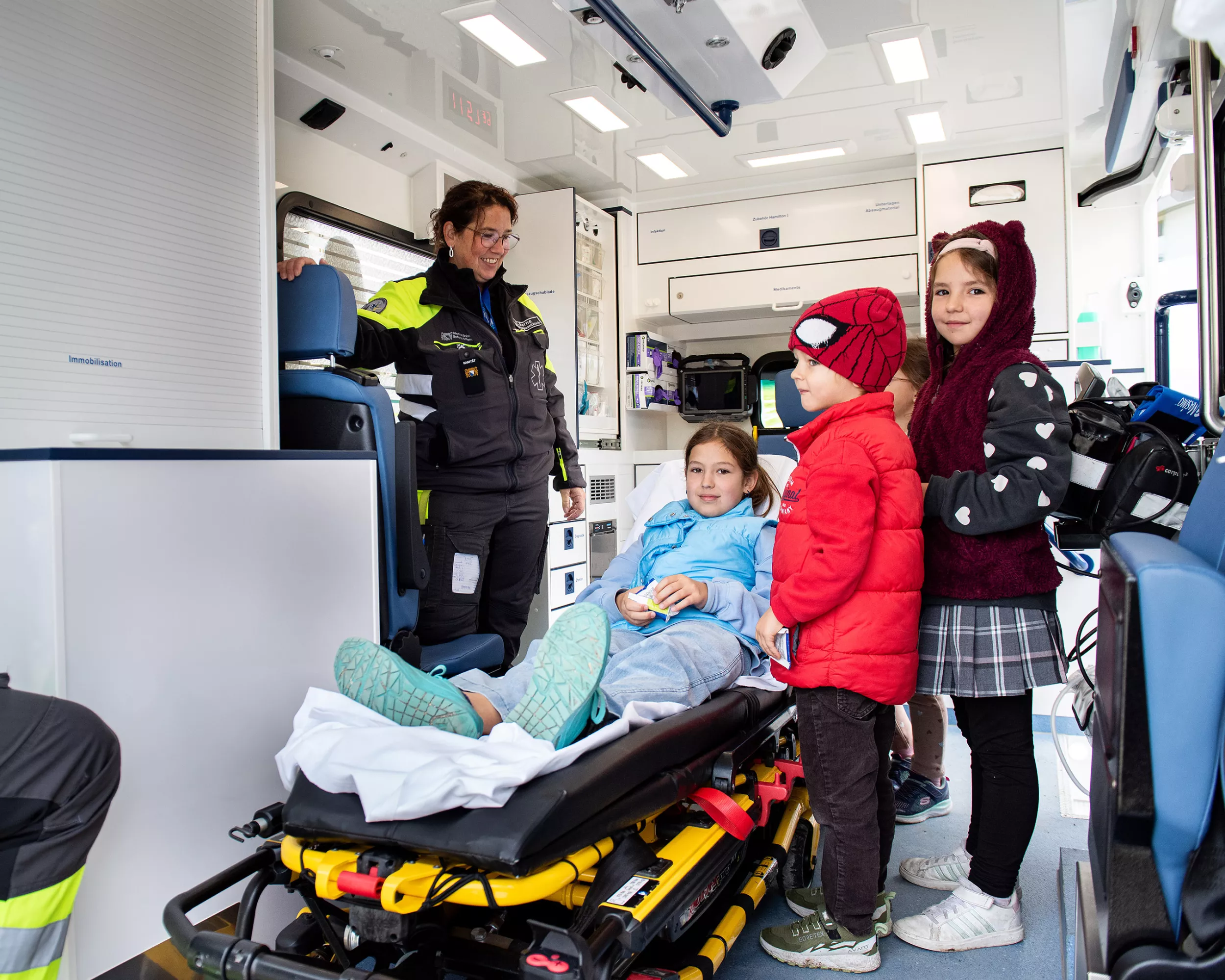 Eine Sanitäterin zeigt drei Kindern den Innenraum eines Rettungswagens. Ein Kind liegt auf einem mobilen Bett.