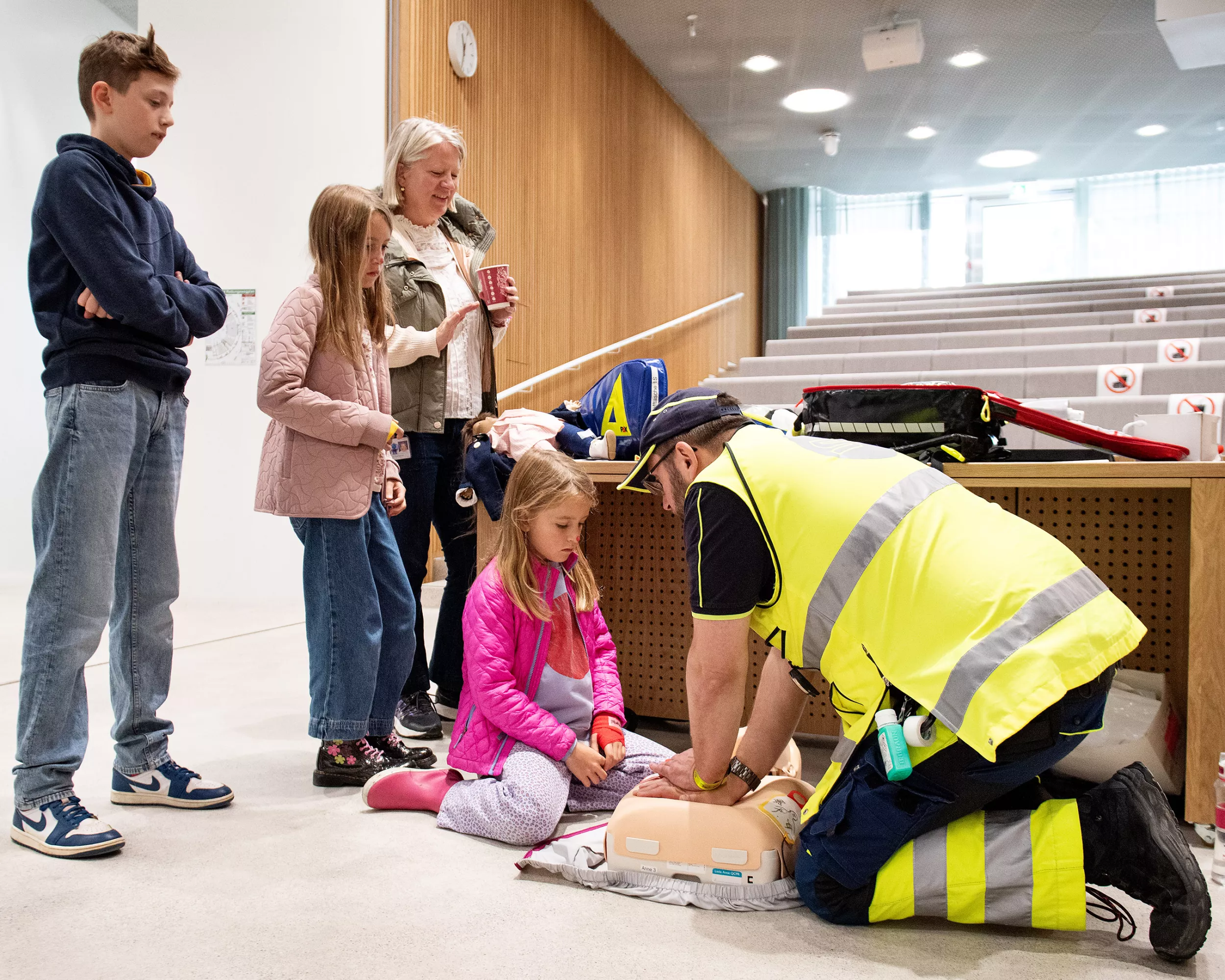 Ein Mann in Uniform demonstriert Reanimation an einer Puppe für eine Gruppe von Kindern und Erwachsenen.