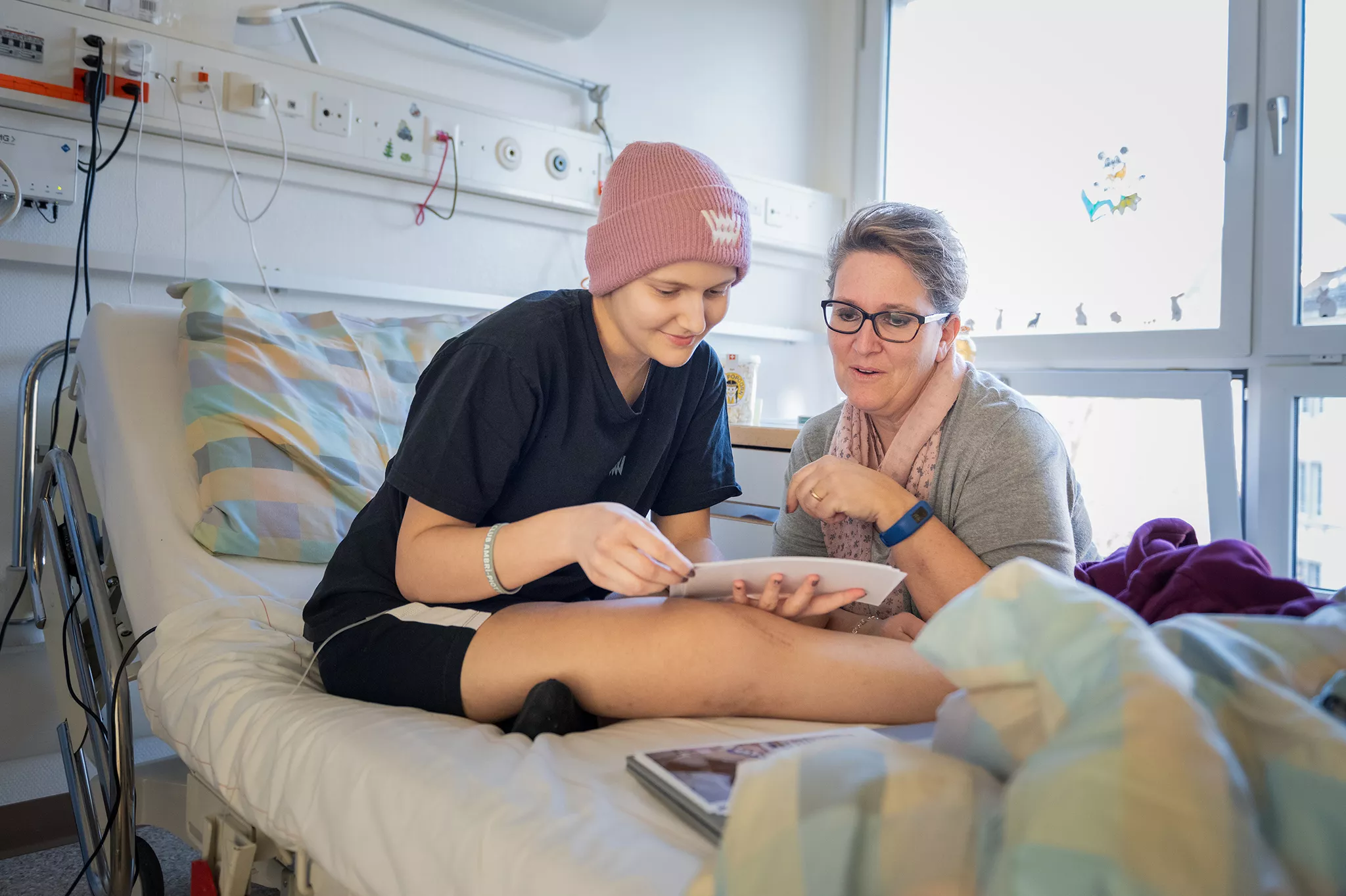 Eine Onkologiepatientin sitzt auf dem Bett. Sie trägt eine Mütze, T-Shirt und Shorts. Neben ihr sitzt ihre Mutter. 