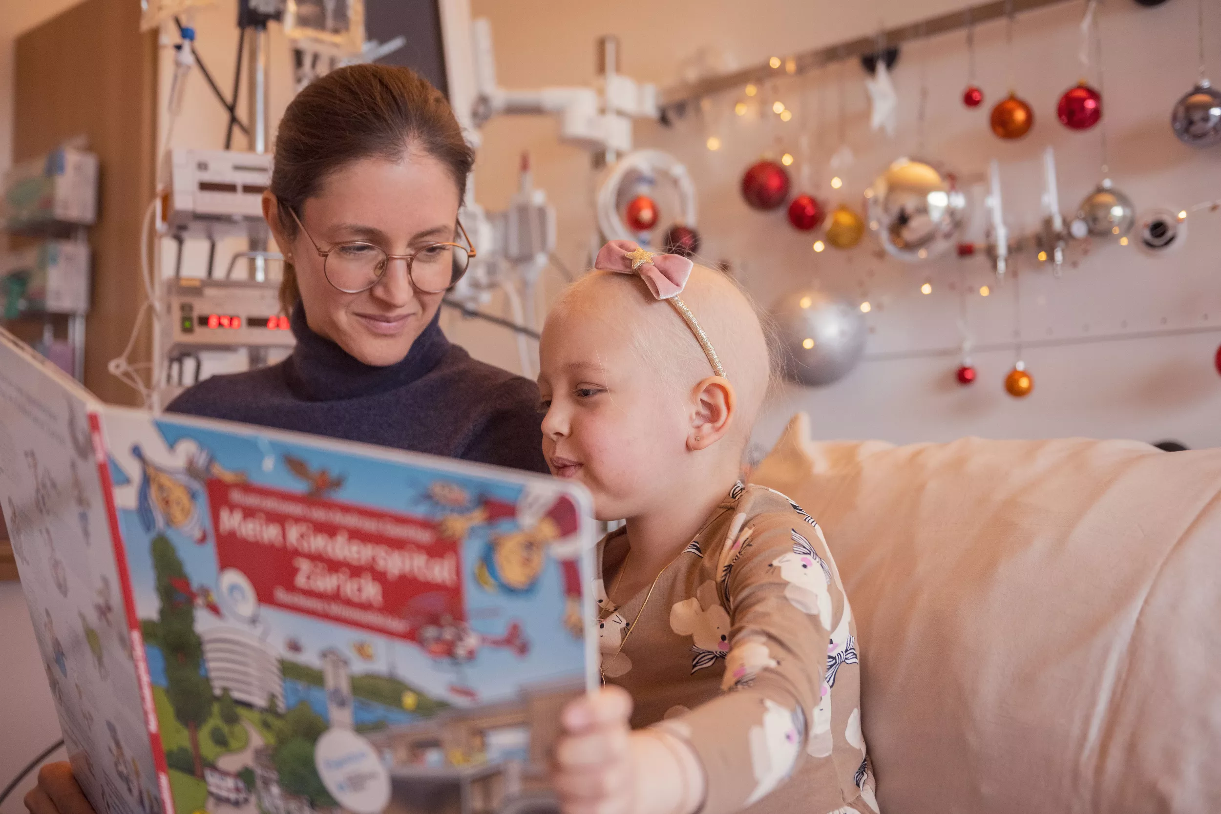 Eine junge Onkologie-Patientin schaut mit ihrer Mutter im Spitalbett ein Wimmelbuch an.
