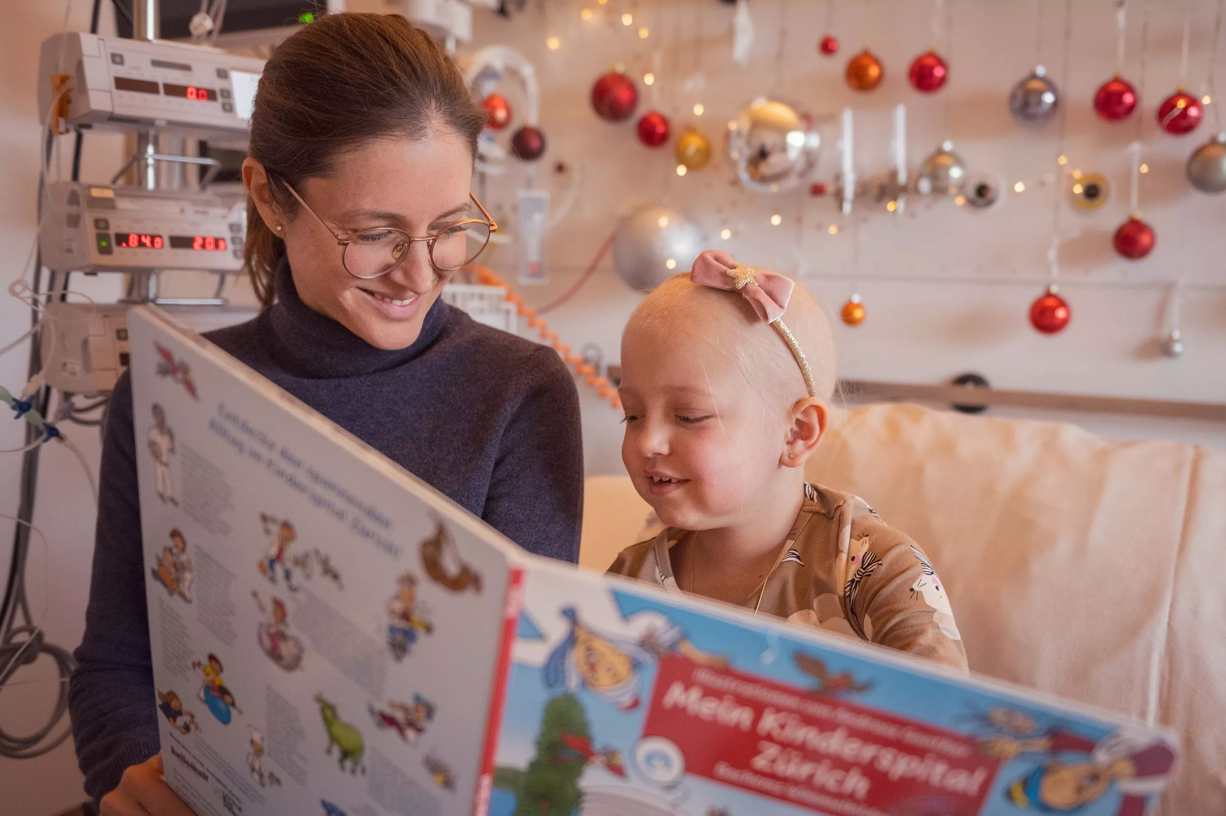Eine Mutter und ein Kind sitzen auf einem Spitalbett und schauen ein Buch an. Der Raum ist weihnachtlich geschmückt.