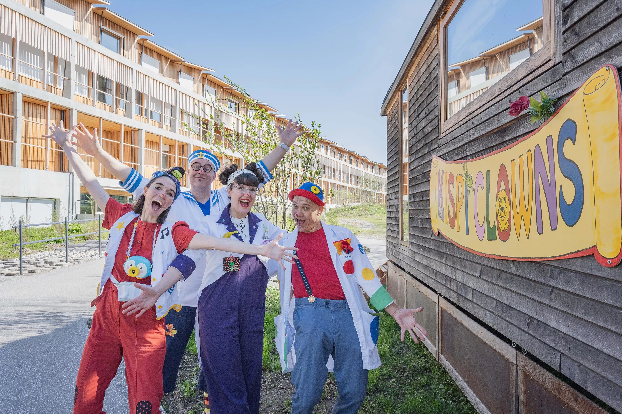 Die vier Spitalclowns des Kinderspitals stehen neben ihrem Zirkuswagen, im Hintergrund ist die Fassade des Kinderspitals
