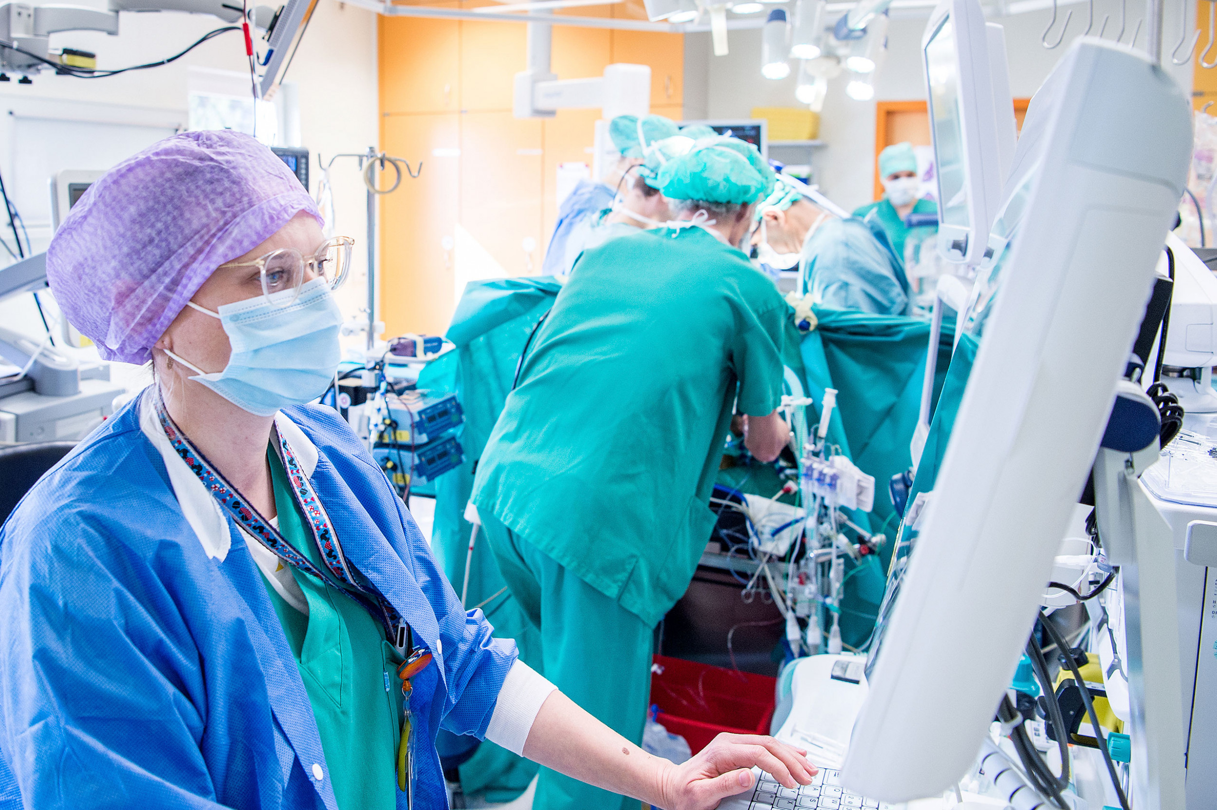 Anästhesistin ist im Herzop am Computer beschäftigt, im Hintergrund OP-Tisch mit Chirurgen