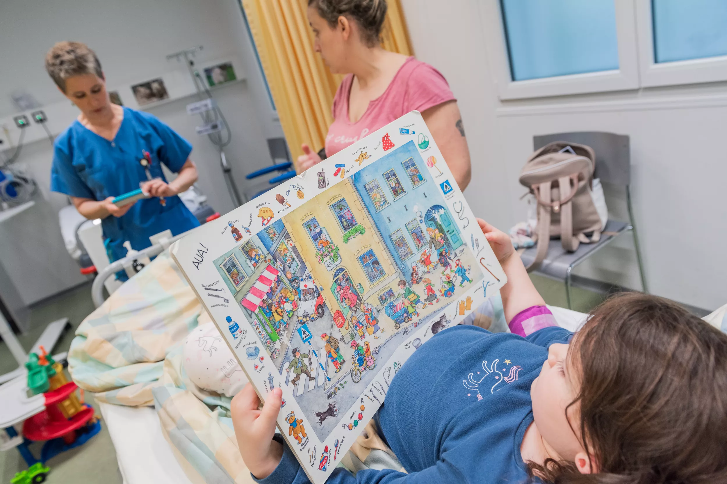 Eine Rheumapatientin liegt auf einem Spitalbett. In ihren Händen hält sie ein Kinderbuch mit einer grossen Zeichnung in der Mitte. Aussenrum sieht man verschiedene Gegenstände mit ihren Bezeichnungen. 
