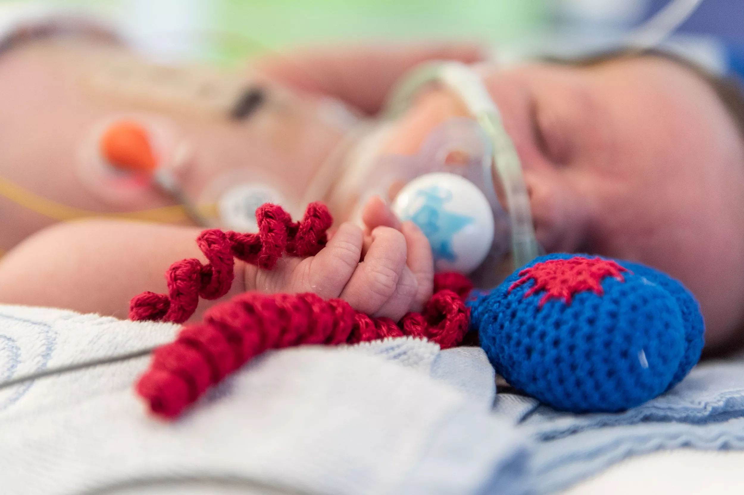 Ein neugeborenes Kind auf der Neonatologie hält ein gestricktes Herz in er Hand um den Greifreflex zu unterstützen