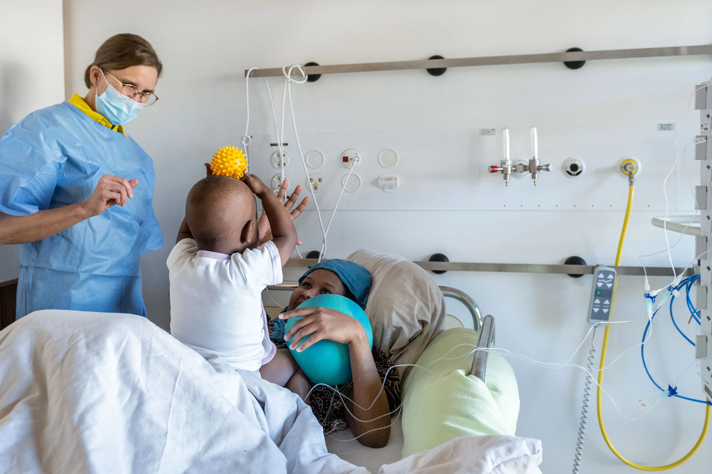 Psychomotorik-Therapie, Patient und Therapeutin spielen mit gelbem Ball, im Hintergrund die Patientenmutter