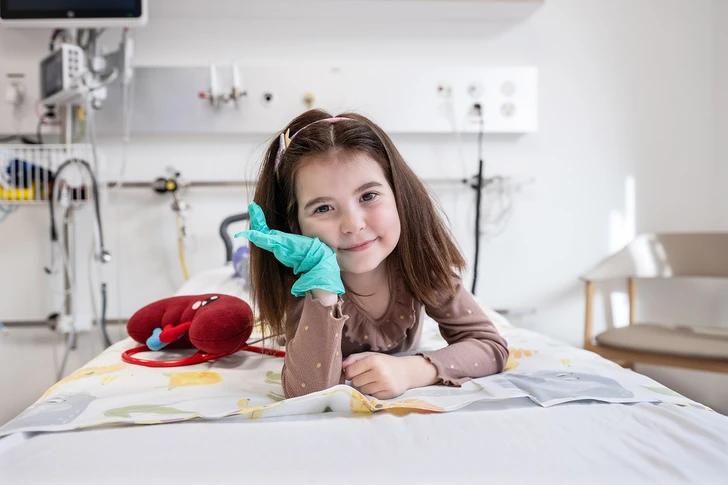 Nephrologie-Patientin Naila liegt auf einem Spitalbett im Kinderspital Zürich. Neben ihr liegt eine Plüsch-Niere und sie trägt einen grünen Gummihandschuh. Sie lächelt in die Kamera.